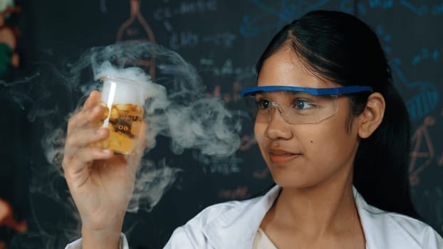 Closeup of smart highschool student inspect colored solution at blackboard. Beautiful scientist holding beaker and looking at chemical liquid while standing in front of blackboard at lab. Edification.