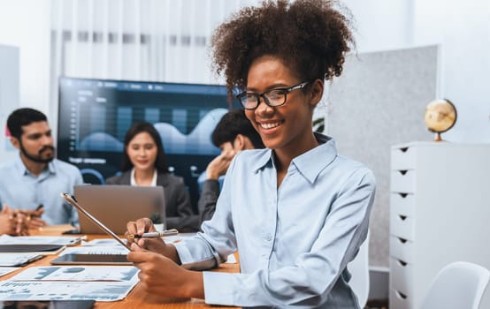 Happy young african businesswoman wearing glasses portrait with group of office worker on meeting with screen display business dashboard in background. Confident office lady at team meeting. Concord