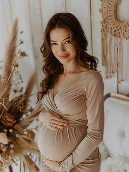 A pregnant woman in a beige gown is showcasing her fashion design, gently holding her waist in a room. The dress features a high neckline and long sleeves, emphasizing her beautiful baby bump