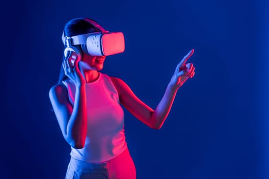 Smart female stand with surrounded by cyberpunk neon light wear VR headset connecting metaverse, futuristic cyberspace community technology. Woman using finger pointing virtual object. Hallucination.