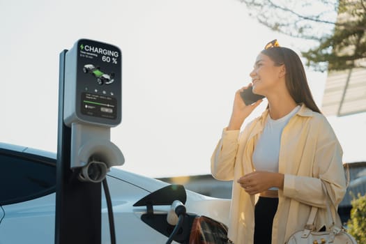 Young woman holding shopping bag talking on the phone while recharging EV car battery from charging station at city mall parking lot. Modern woman go shopping by eco car. Expedient