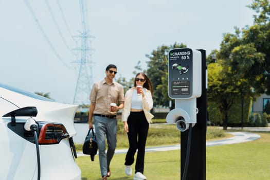 Young couple with coffee recharge EV car battery at charging station connected to electrical industrial power grid tower. Couple with shopping and travel using eco electric car lifestyle. Expedient