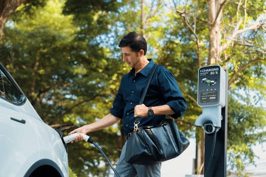 Young man recharge electric car's battery from charging station in outdoor green city park in springtime. Rechargeable EV car for sustainable environmental friendly urban travel lifestyle. Expedient