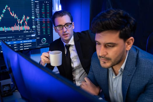 Two stock exchange traders discussing dynamic investment graph in currency rate on monitor at night. Businessman partners coffee meeting in high stock market in neon light at workplace. Sellable.