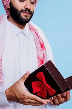 Muslim man wearing traditional cultural attire opening gift box for special occasion. Arab dressed in white thobe holding festive present giftbox with bow ribbon in hands closeup