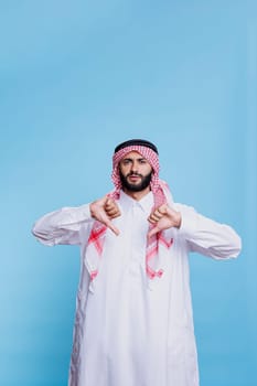 Discontent arab wearing traditional clothes posing with thumbs down and looking at camera with serious expression. Muslim man dressed in thobe and ghutra and showing disapproval gesture