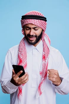 Excited man wearing traditional muslim outfit checking good news message while using smartphone. Happy cheerful muslim person showing winner gesture while scrolling internet page on mobile phone
