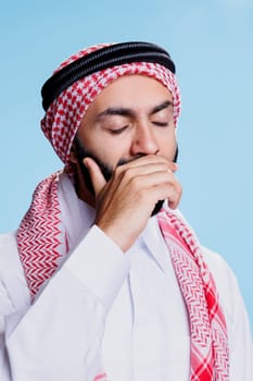 Sleepy man with closed eyes wearing traditional muslim headdress and thobe yawning. Exhausted arab dressed in islamic clothes covering mouth with arm while feeling sleepiness and tiredness