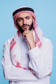 Muslim man with in traditional attire contemplating with pensive look. Questioned arab person wearing thobe and ghutra headscarf posing in thinking pose with hand supporting chin