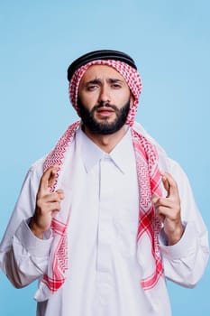 Muslim man dressed in traditional clothes making wish with crossed fingers and looking at camera with hopeful expression. Arab wearing thobe praying for luck with superstitious gesture portrait