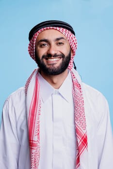Cheerful muslim man dressed in islamic white thobe and checkered ghutra headdress studio portrait. Happy arab person standing and looking at camera with carefree expression