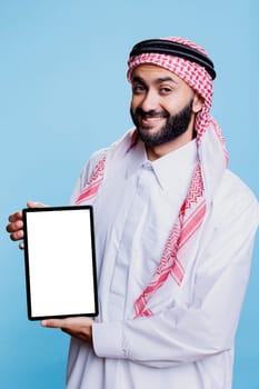 Smiling arab dressed in traditional attire holding digital tablet with white blank screen mockup and looking at camera. Muslim man promoting portable gadget empty touchscreen for application ads