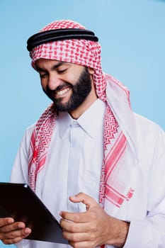 Arab man laughing while enjoying internet entertainment activity on digital tablet. Smiling muslim person having fun while chatting online and scrolling social media on gadget touchscreen