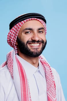 Smiling arab man wearing traditional headscarf looking at camera with cheerful face expression. Joyful muslim person in ghutra headdress posing for studio portrait on blue background