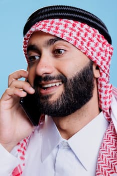 Excited muslim man wearing traditional checkered headscarf answering smartphone call with smiling expression. Cheerful arab dressed in islamic headdress speaking on mobile phone closeup