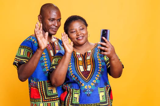 Smiling african american man and woman wearing ethnic clothes waving hi while speaking in smartphone videocall. Cheerful couple showing greeting gesture while having online communication on phone