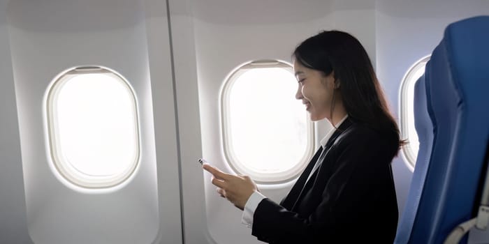 Asian young businesswoman successful or female entrepreneur in formal suit in a plane sit in a business class seat and uses a smartphone during flight. Traveling and Business concept.