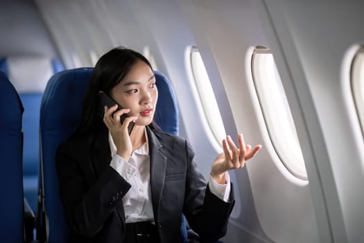 Asian young businesswoman successful or female entrepreneur in formal suit in a plane sit in a business class seat and uses a smartphone during flight. Traveling and Business concept.