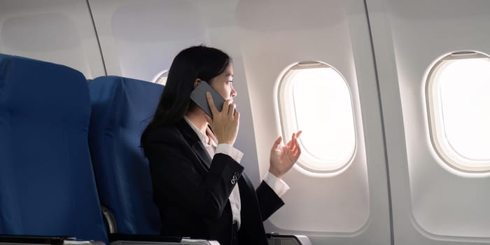 Asian young businesswoman successful or female entrepreneur in formal suit in a plane sit in a business class seat and uses a smartphone during flight. Traveling and Business concept.