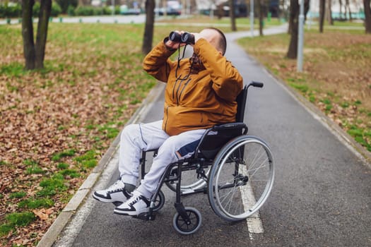 Paraplegic handicapped man in wheelchair is using binoculars outdoor. He is watching birds.