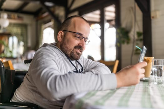 Happy paraplegic handicapped man in wheelchair is sitting at restaurant and using smartphone.