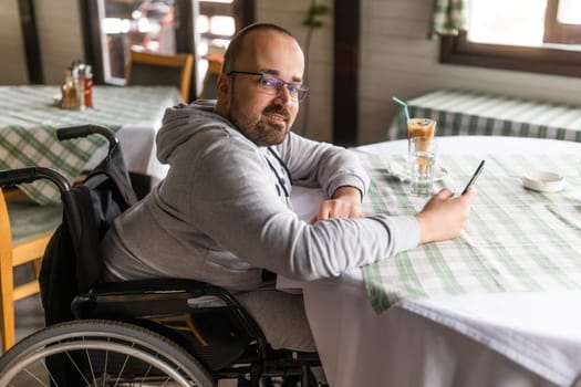 Happy paraplegic handicapped man in wheelchair is sitting at restaurant and using smartphone.