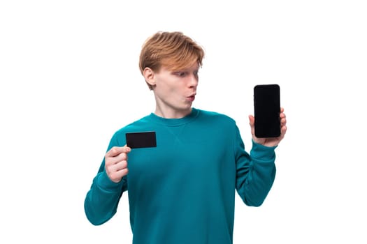 young caucasian student guy with red hair dressed in a blue sweater holds a plastic card mockup and a smartphone.