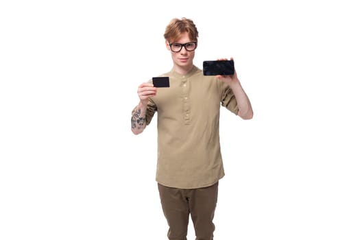 handsome young caucasian male promoter with red hair and a tattoo on his arm dressed in a khaki shirt holds a credit card and a phone.