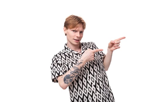 a young handsome guy with red hair and a tattoo on his arms, dressed in a short-sleeved shirt, points with his hand to the space for advertising. business concept.