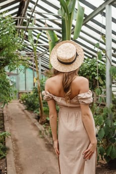 A beautiful young woman takes care of plants in a greenhouse. The view from the back. Concept of gardening and an eco-friendly lifestyle.