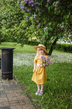 A little girl in a yellow dress and straw hat wearing a bouquet of lilacs. A walk in a spring park, blossoming lilacs