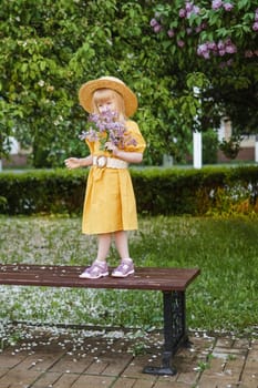 A little girl in a yellow dress and straw hat wearing a bouquet of lilacs. A walk in a spring park, blossoming lilacs