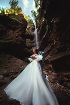 Couple in love against the backdrop of a waterfall. Honeymoon trip. Happy couple in the mountains. Vertical photo of newlyweds hugging against the backdrop of a waterfall in the gorge.