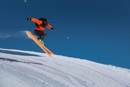A skier in an orange jacket and a black helmet makes a jump on a sunny day against a clear sky. Copy space for your information.