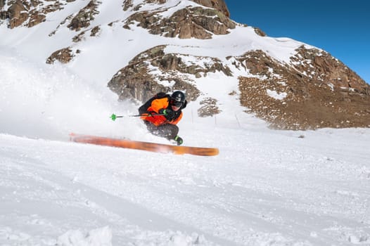 Pro skier in an orange jacket and a black helmet with a mask rides down a snowy slope at high speed. Powder from under skis and a look at the camera
