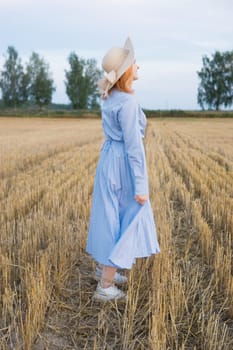 A red-haired woman in a hat and a blue dress walks in a field with haystacks. The view from the back