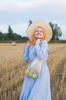 A red-haired woman in a hat and a blue dress walks in a field with haystacks