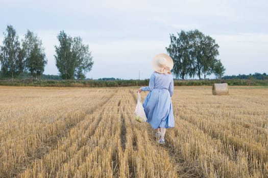 A red-haired woman in a hat and a blue dress walks in a field with haystacks. The view from the back