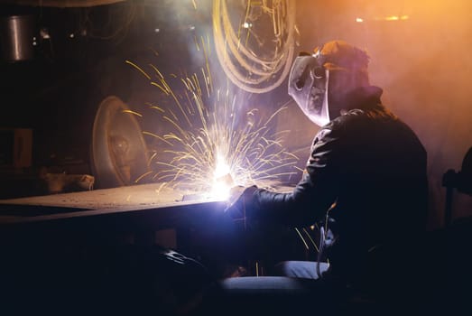 Young mechanic with a welding machine in an old dirty garage at night. Man in protection doing welding work, hobby.