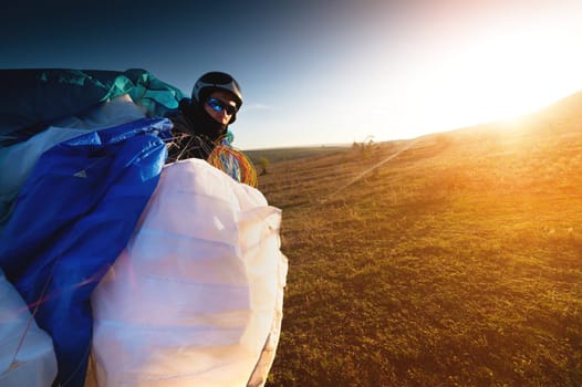 A young male paraglider at sunset in the mountains collects his paraglider to take it to the starting point. Paragliding sport.