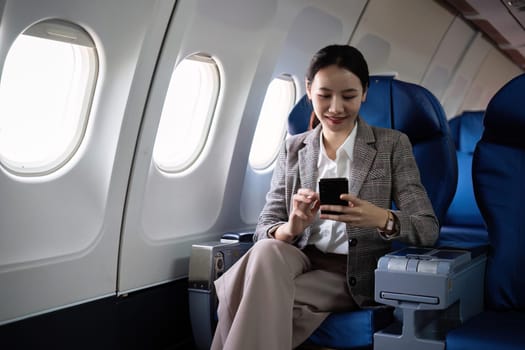 Young Asian woman checks business news on mobile phone, sitting near window in first class on airplane during flight, travel and business concept.