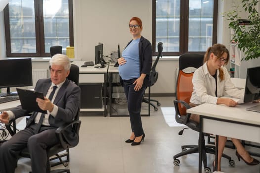 A happy pregnant woman stands in the middle of the office next to a Caucasian woman and an elderly man working at computers