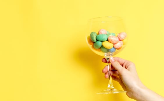 The hand of a young Caucasian unrecognizable girl holds a wine glass with Easter and marble decorative eggs on the right on a yellow background with copy space on the left, side view close-up.