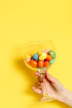 The hand of a young Caucasian unrecognizable girl holds a tilted wine glass with Easter marble decorative eggs on a yellow background with copy space from above, side view close-up.