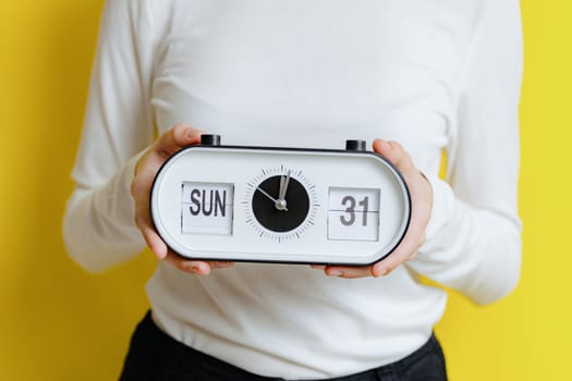 The hands of one young Caucasian unrecognizable girl holds a clock with time, date and day: Sunday, lunch, 31, standing on a yellow background on a spring day in the room, side view close-up.
