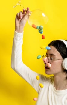 Portrait of one young Caucasian beautiful happy teenage girl with closed eyes in glasses and headphones pours out of a wine glass with Easter marble decorative eggs onto her face, sitting sideways on a yellow background on a spring day in the room, side view close-up.