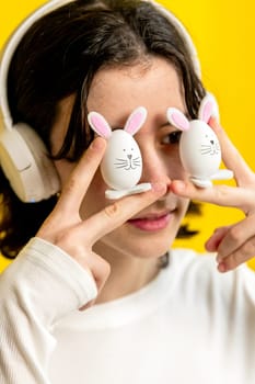 Portrait of one young Caucasian beautiful happy teenage girl holding with her fingers two Easter decorative bunny at eye level, standing half-turned on a yellow background on a spring day in the room, side view close-up with depth of field.