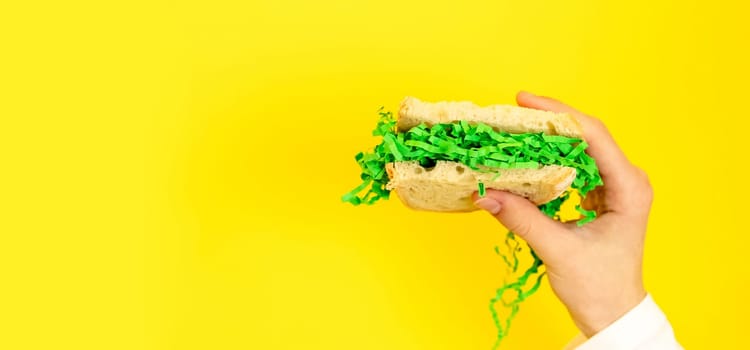 Portrait of one hand of a young Caucasian teenage girl holding a bread sandwich on the right with Easter decorative green hay on a yellow background with with copy space on the left on a spring day in the room, side view close-up with depth of field.