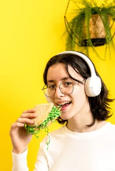 Portrait of one young Caucasian beautiful happy teenage girl holding, about to eat a bread sandwich with Easter decorative green hay, standing on a yellow background with a hanging palm flower on a spring day in the room, side view close-up with selective focus.