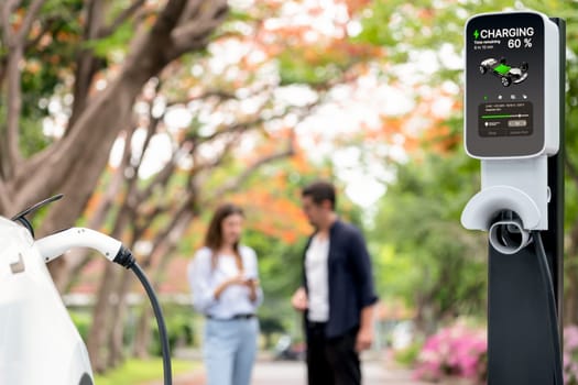 Focused EV car recharging battery on blurred background of lovey couple during autumnal road trip travel with electric vehicle recharging battery. Eco friendly travel on vacation during autumn. Exalt